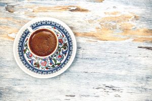 A photo taken from above of a cup of Turkish or Greek coffee on a decorative sauce sitting on a white wooden background.