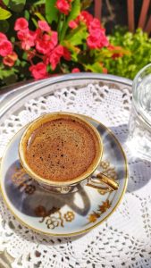 A cup of Arabic or Greek coffee sits on a decorative saucer on a tray in a garden in front of deep pink flowers.