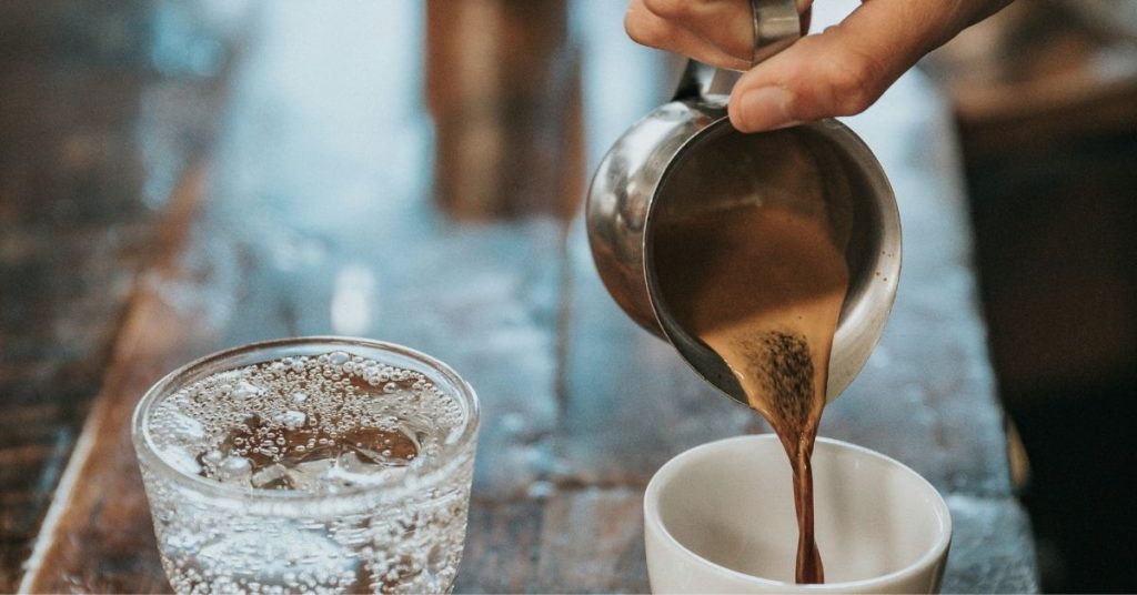 A hand pours Arabic coffee from a metallic jug into a white mug sitting on a wooden table next to a glass of carbonated water.