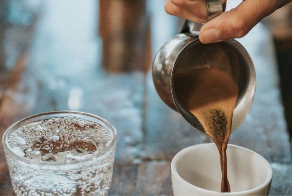 A hand pours Arabic coffee from a metallic jug into a white mug sitting on a wooden table next to a glass of carbonated water.