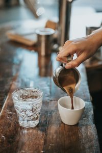A hand pours Arabic coffee from a metallic jug into a white mug sitting on a wooden table next to a glass of carbonated water.